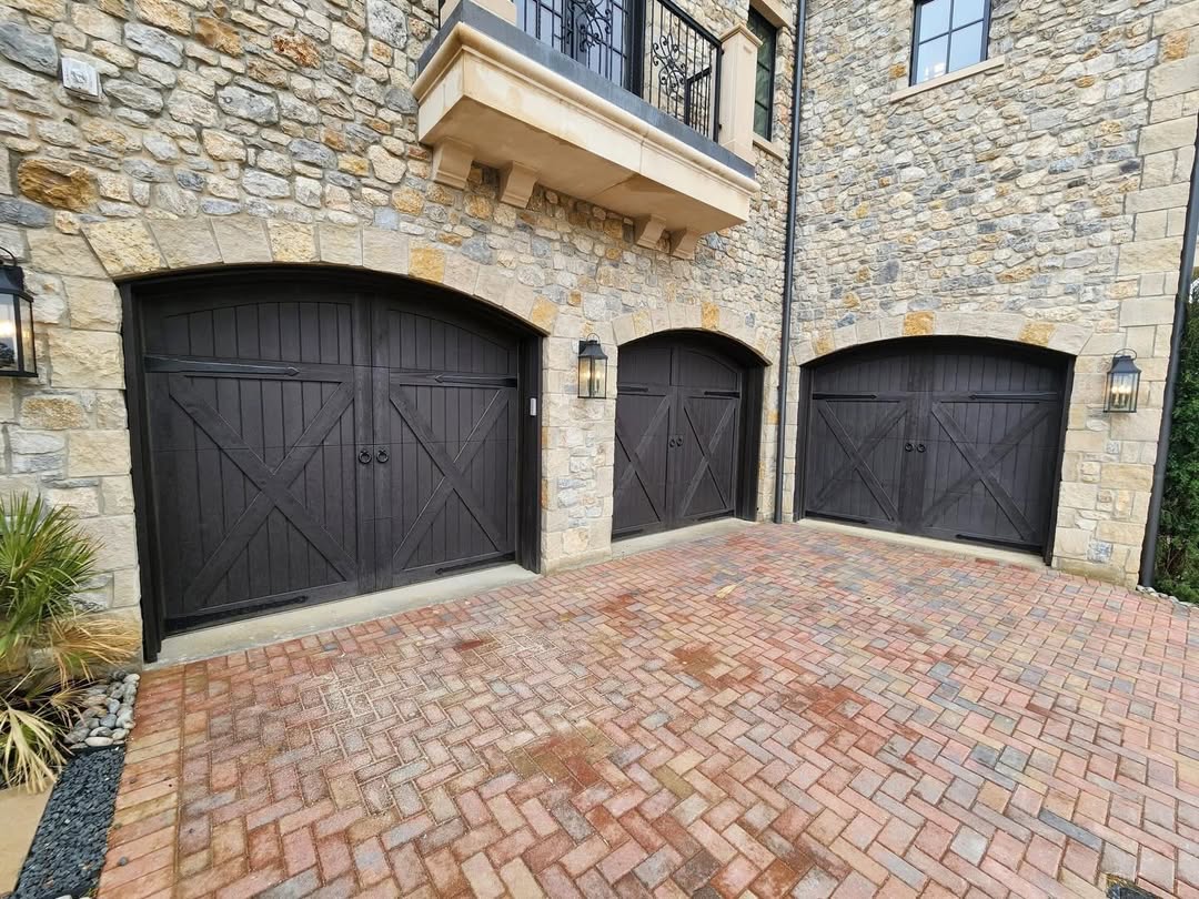 Three espresso arched X-brace custom wood garage doors on a stone home